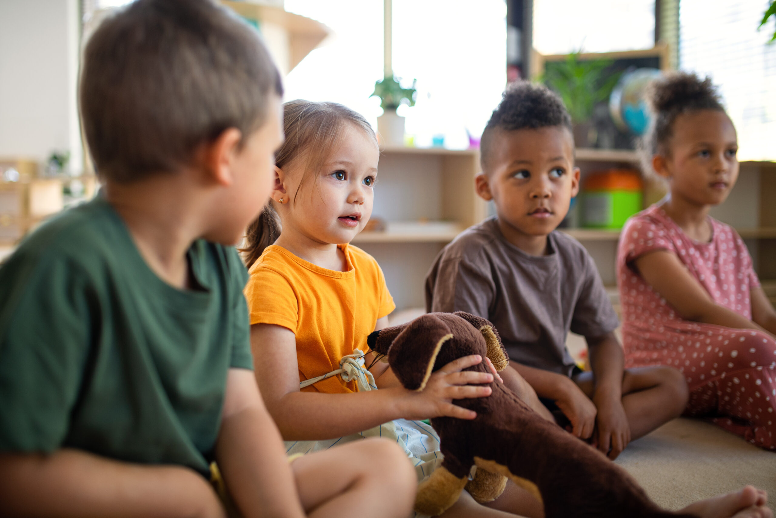 Group of small nursery school children sitting on floor indoors in classroom, listening to teacher.